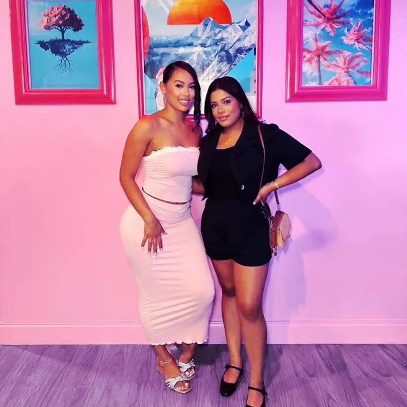 Two smiling women pose in front of a pink wall with colorful framed artwork.