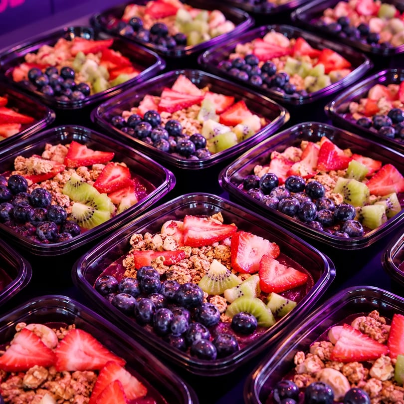 Rows of acai bowls topped with granola, blueberries, strawberries, and kiwi in clear containers.