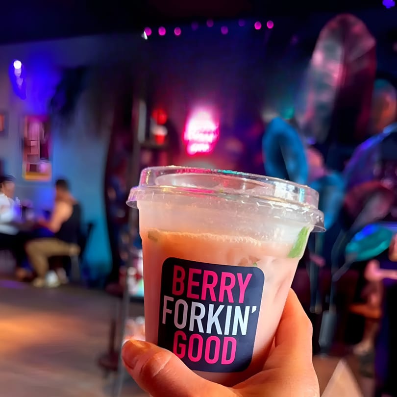 Hand holding a pink drink with 'BERRY FORKIN' GOOD' label in a vibrant, dimly lit restaurant.