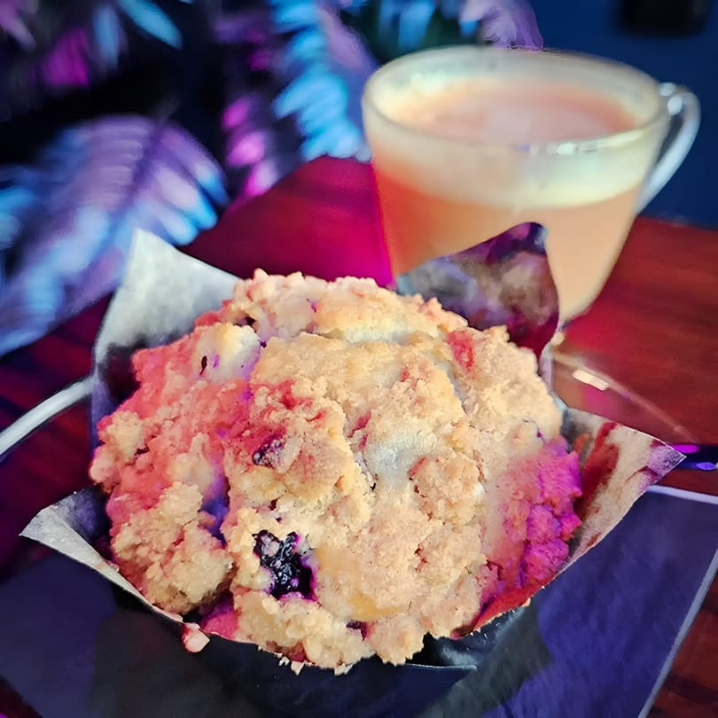 Blueberry muffin with crumb topping and a latte in a clear glass, against a colorful, blurry background.