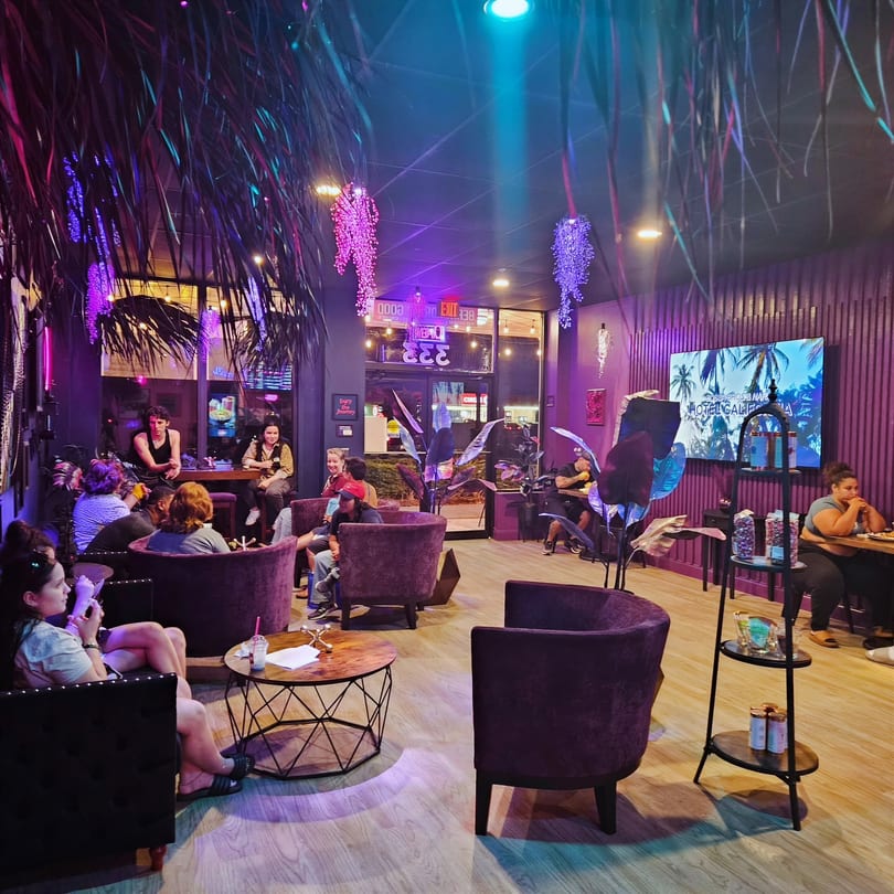 People relaxing in a vibrant lounge with purple and blue lighting, comfortable armchairs, and tropical decor.