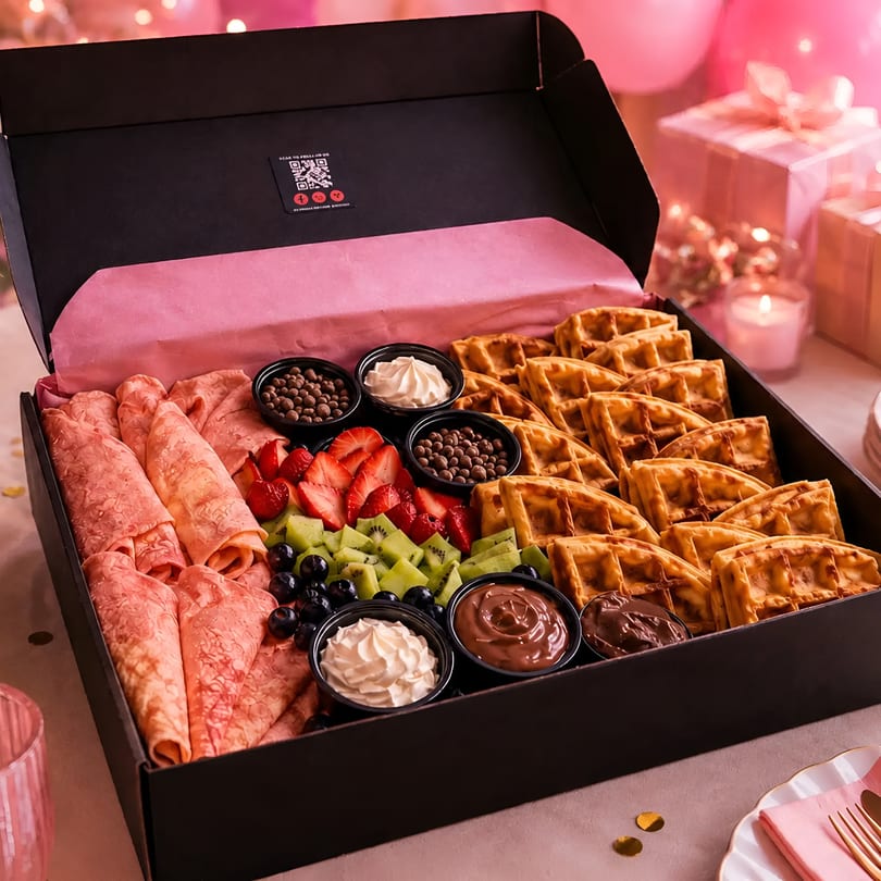 Gourmet breakfast box with mini waffles, rolled crepes, fresh fruit, and sweet dips on a pink party table.
