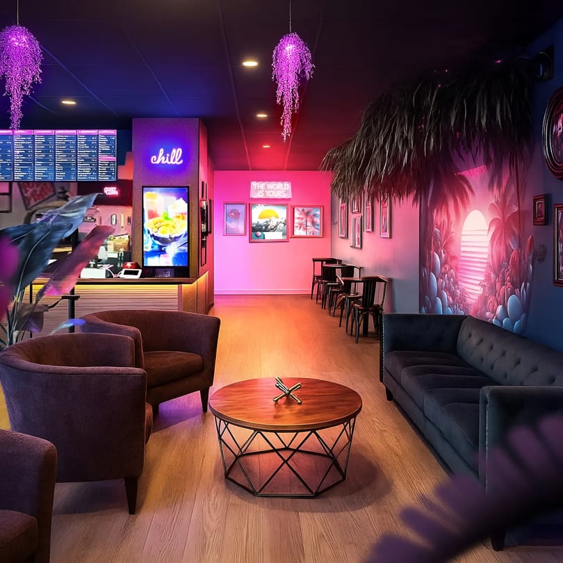 Modern restaurant interior with lounge seating, a counter, neon signs, and atmospheric pink and purple lighting.