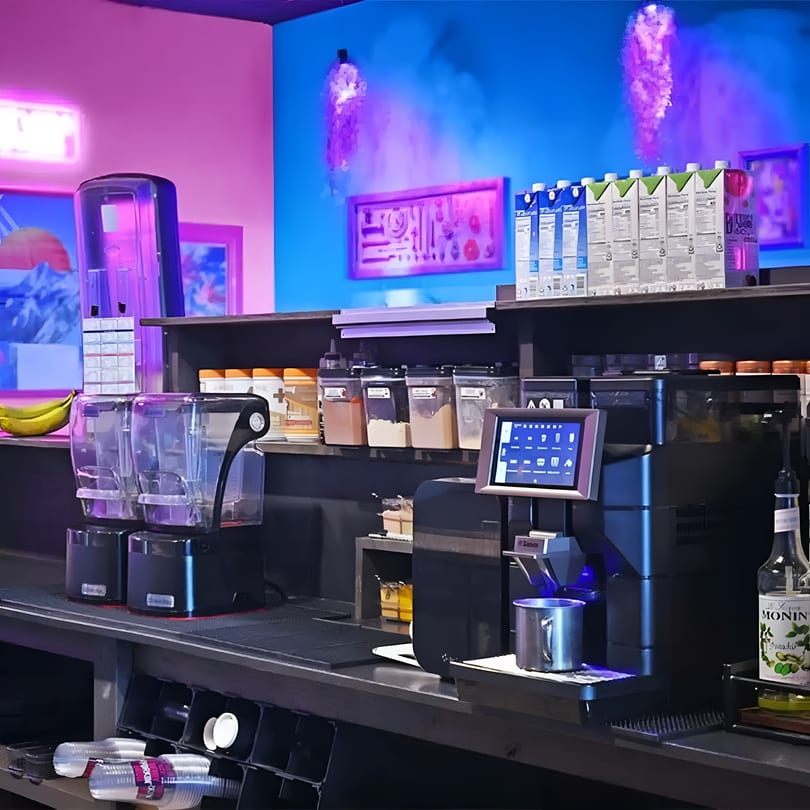 Vibrant cafe counter with blenders, coffee machine, ingredients, and neon pink and blue lighting.