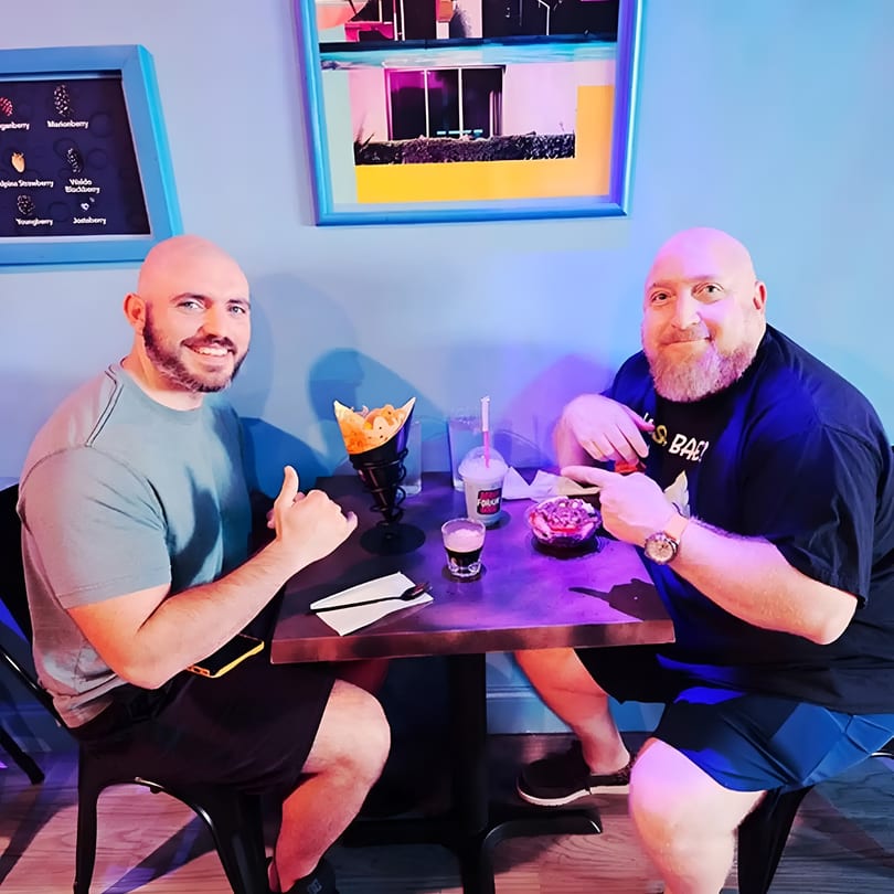 Two smiling men enjoy desserts and drinks at a vibrant restaurant table.