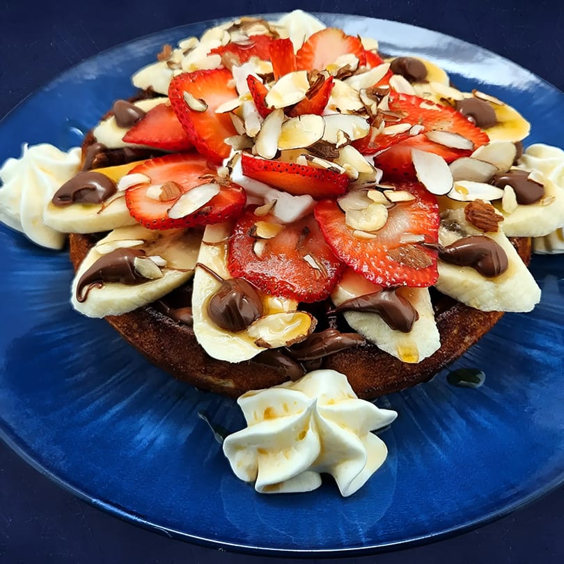 Delicious waffle with strawberries, bananas, chocolate drizzle, whipped cream, and almonds on a blue plate.