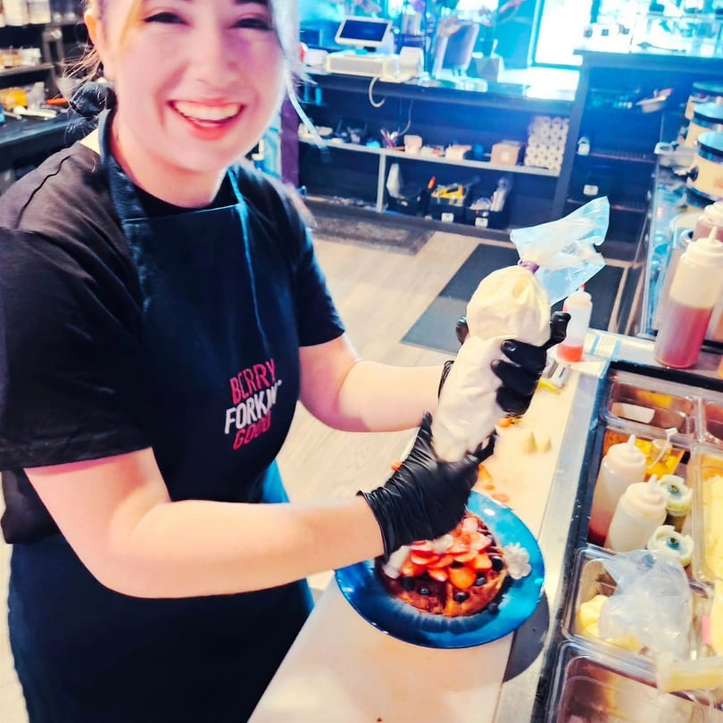 Smiling woman in a "BERRY FORK" apron piping whipped cream onto a berry waffle.