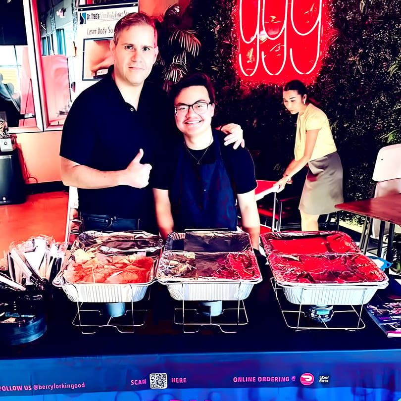 Two men pose behind a buffet table with three foil food pans, a woman works nearby.