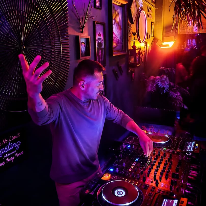 A DJ with raised hand mixing music on a console in a vibrant, neon-lit club.
