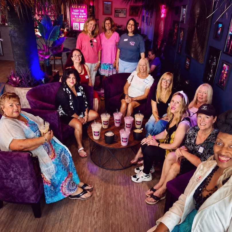 Smiling women enjoy drinks in a stylish lounge with purple furniture and neon lighting.