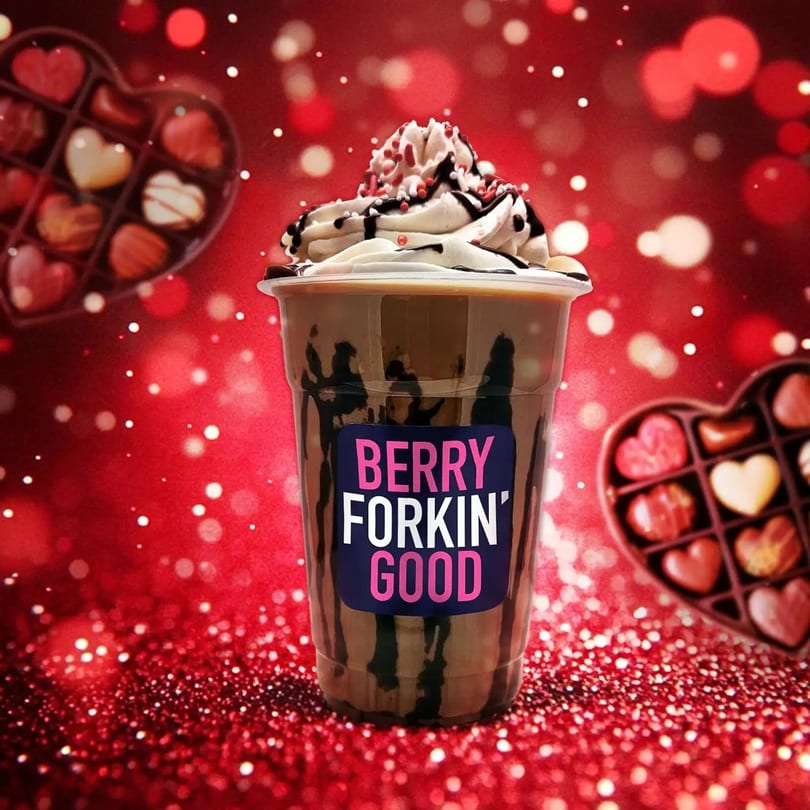 Chocolate drink with whipped cream and sprinkles, labeled 'BERRY FORKIN' GOOD', against a red, glittery background with heart chocolates.